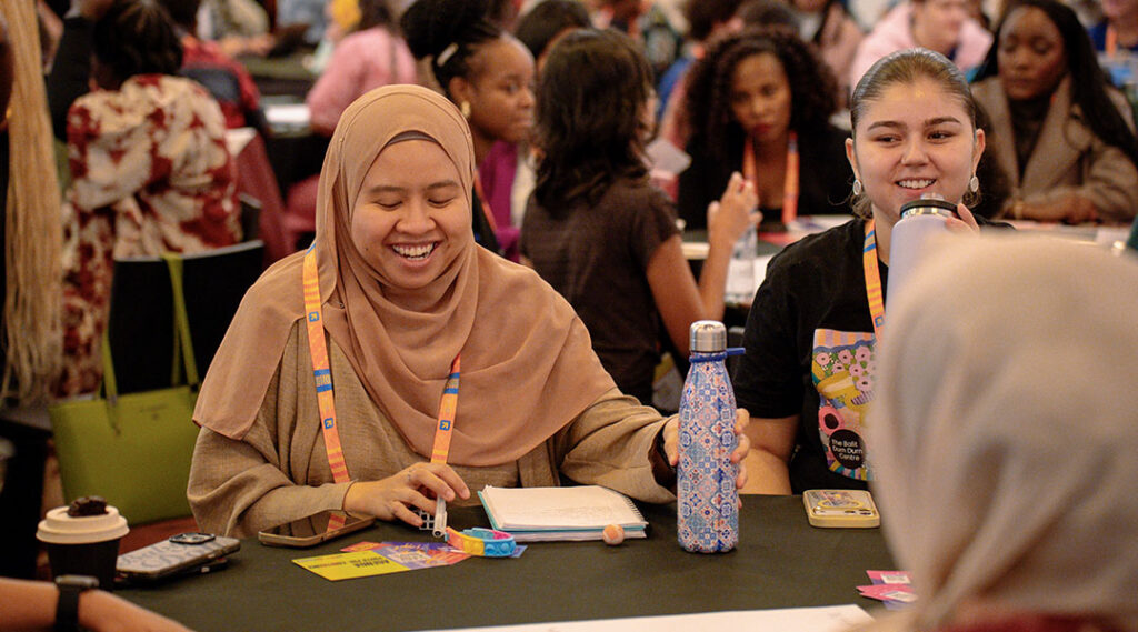Two delegates smile at a workshop table with notebooks, water bottles, and other participants in the background.