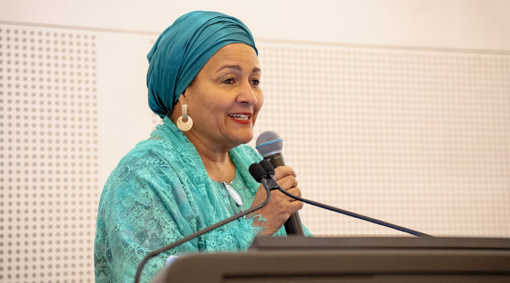Ms. Amina J. Mohammed stands at a podium and speaks into a handheld microphone, smiling, against a light-colored perforated backdrop.