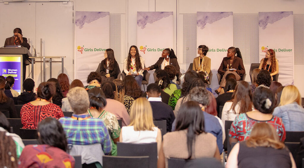 A wide conference-room shot shows a moderator at a lectern and several young panelists seated on stage in front of Girls Deliver banners, with attendees filling the room.