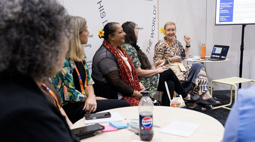 Grupo de participantes asiste a una mesa redonda durante un taller en el Espacio de la Declaración, mientras la persona encargada de facilitarlo interviene junto a una pantalla que reza «objetivos».