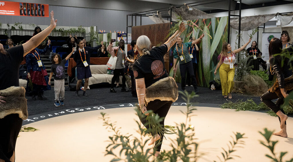Dancers and attendees stand in a circle with raised arms around a decorated indoor gathering space with native plants.