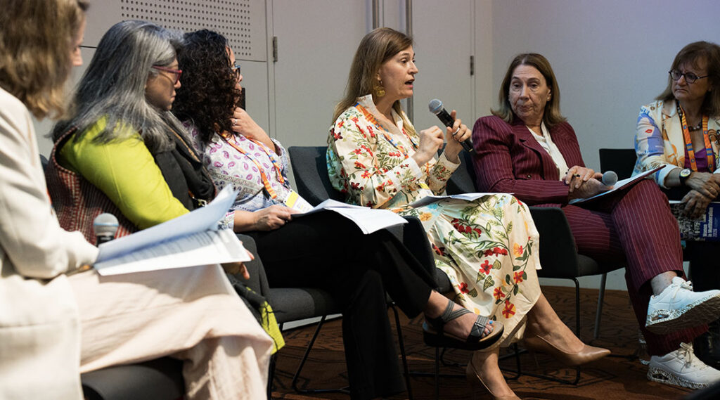 Six panelists sit in a row of chairs; one woman in a floral dress speaks into a handheld microphone.