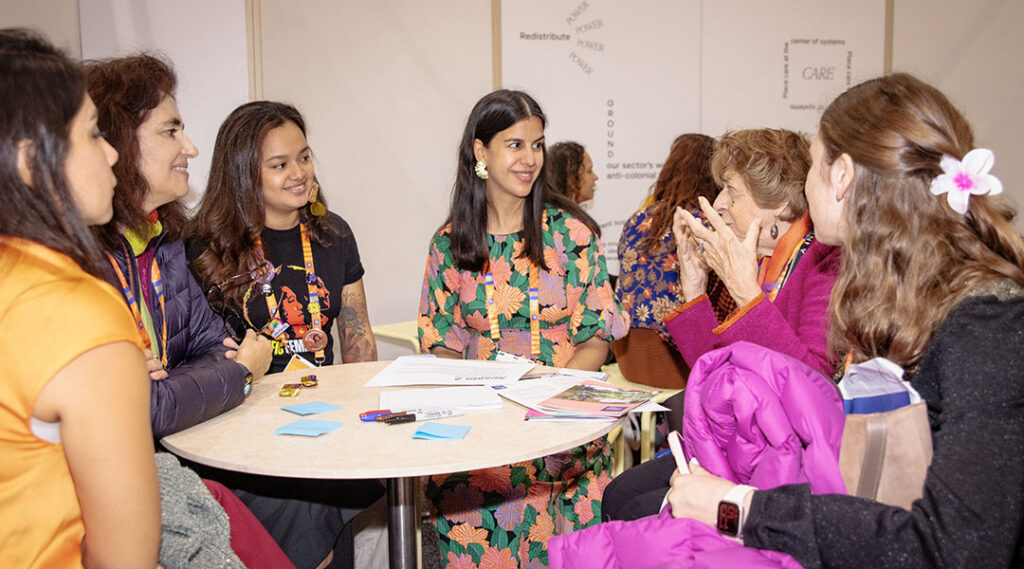 Five people sit around a round table in conversation; behind them, wall text reads 'Redistribute Power' and 'Care' as part of a conference installation.