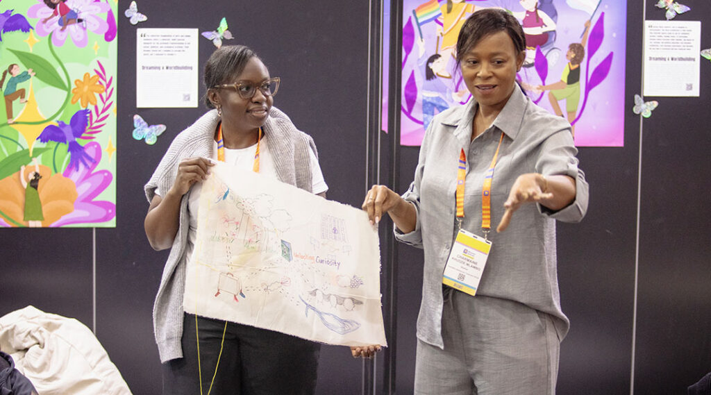 Two women stand holding a hand-drawn artwork on fabric in a Conference space with colorful feminist artwork displayed on the walls behind them.