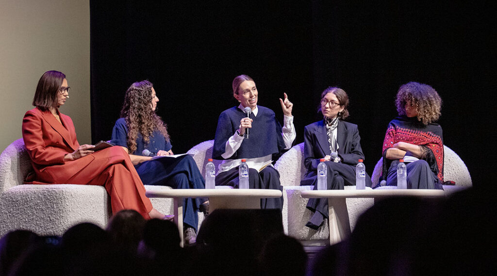 Five panelists sit in white armchairs on a darkened stage; Dame Jacinda Ardern, seated in the center, holds a microphone and gestures while the other panelists listen. The audience is visible in silhouette in the foreground.