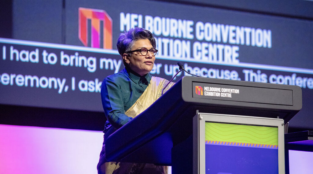 Dr. Maliha Khan stands behind a Women Deliver podium with live captions projected on a large screen behind them.